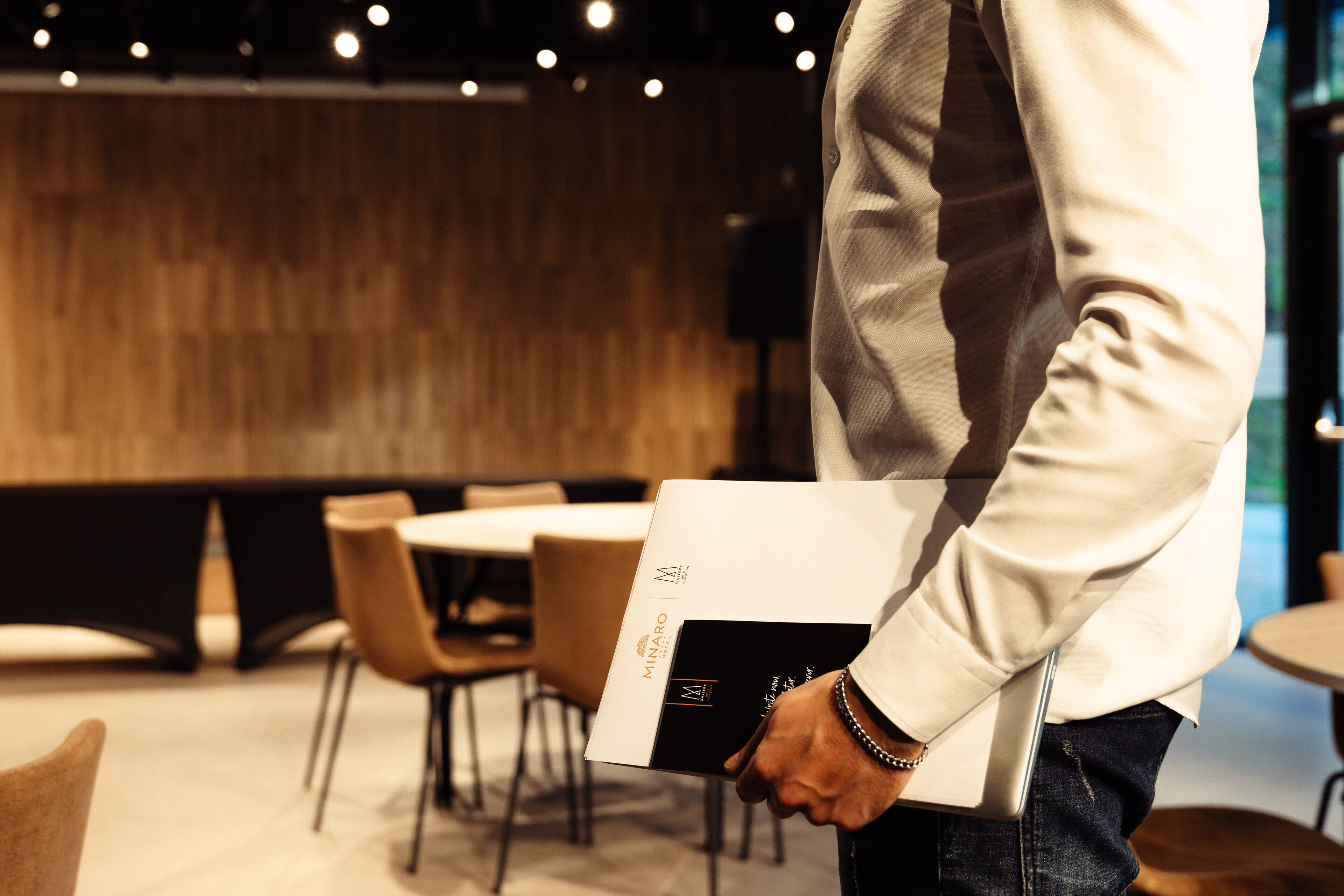 A person holding a book with a logo, standing in a room with tables and chairs.