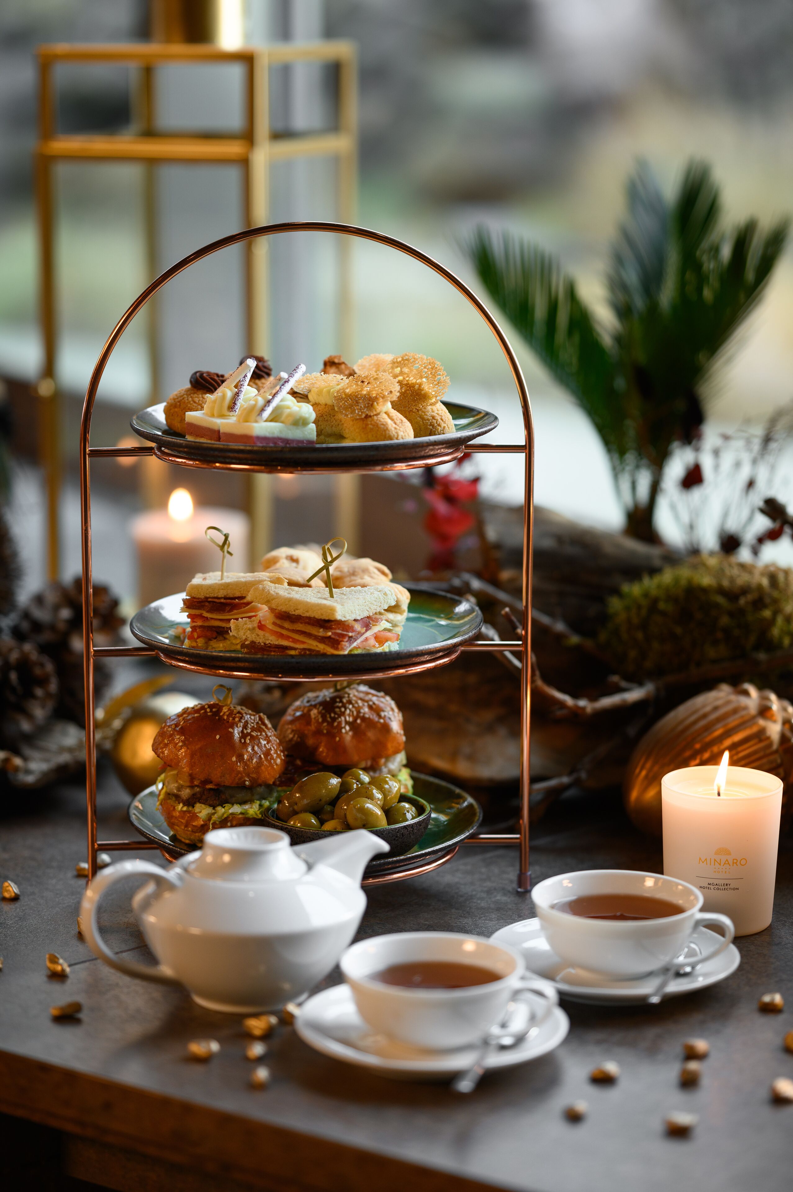 A three-tiered dessert stand with sandwiches and pastries, a teapot, cups, and a candle on a table.