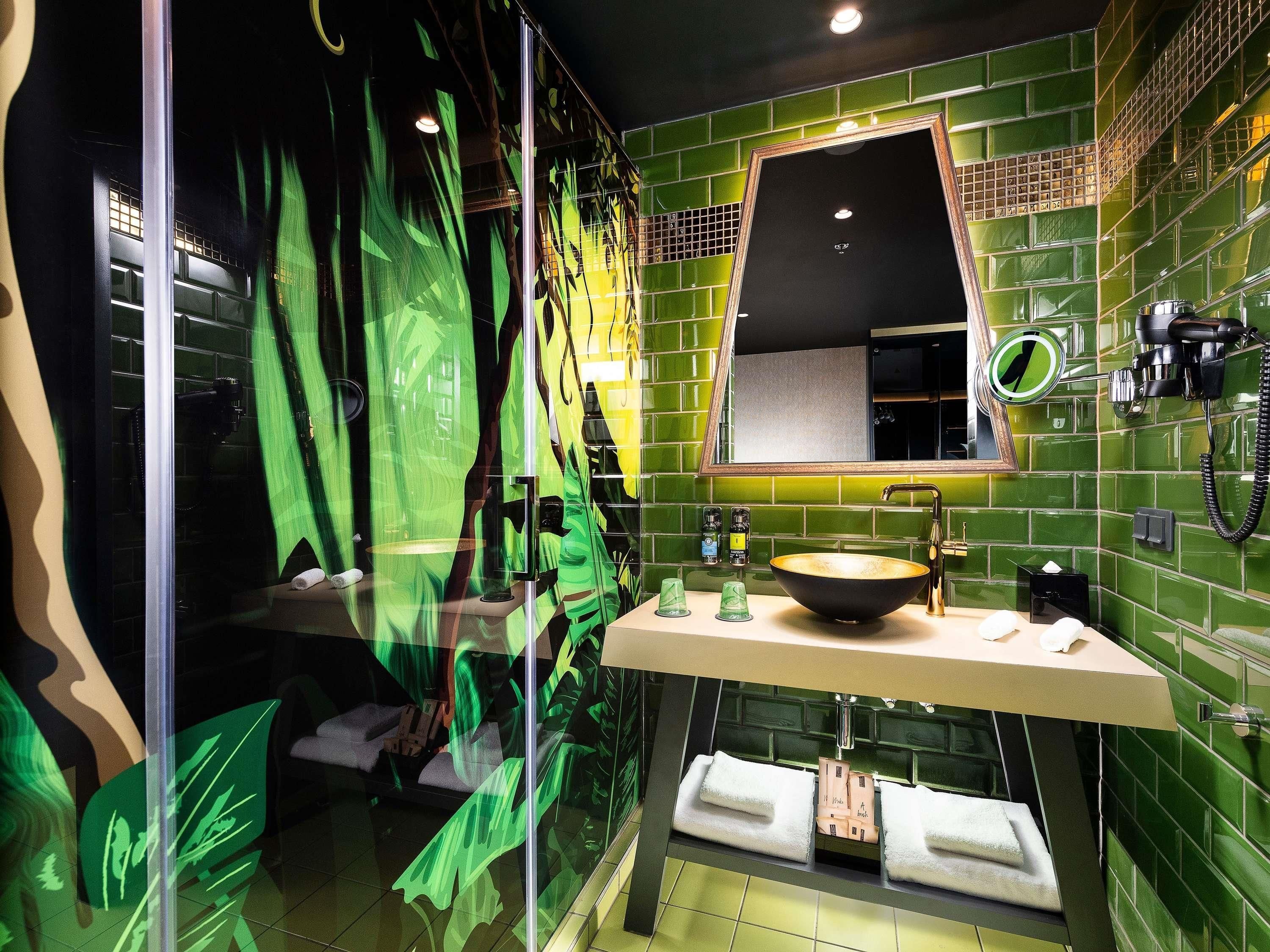 Un baño con pared de azulejos verdes, un espejo grande, un lavabo y un perchero para toallas.