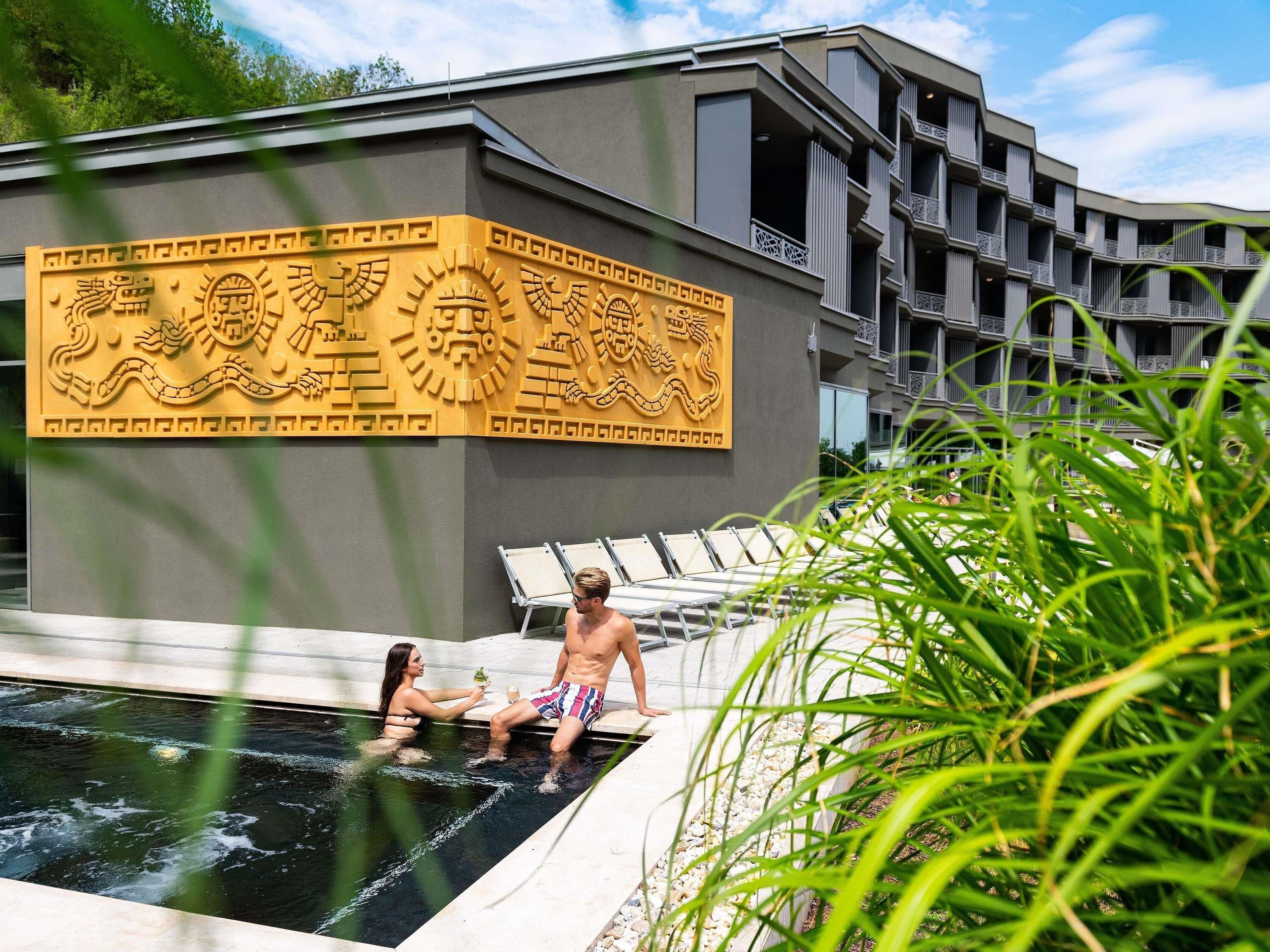 A swimming pool area with a decorative wall, lounge chairs, and a building with balconies.