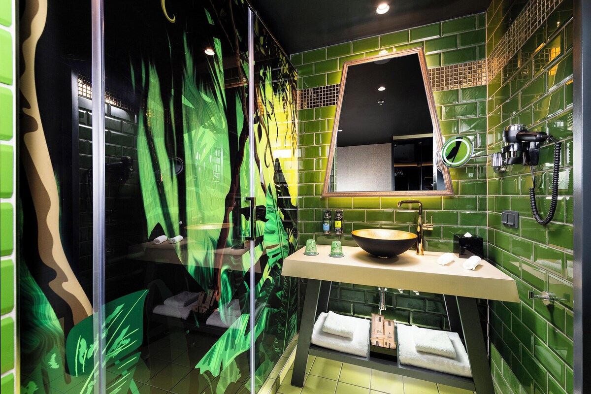 Une salle de bain avec un mur recouvert de carreaux verts, un lavabo avec un receveur, un miroir et un sèche-cheveux fixé au mur.