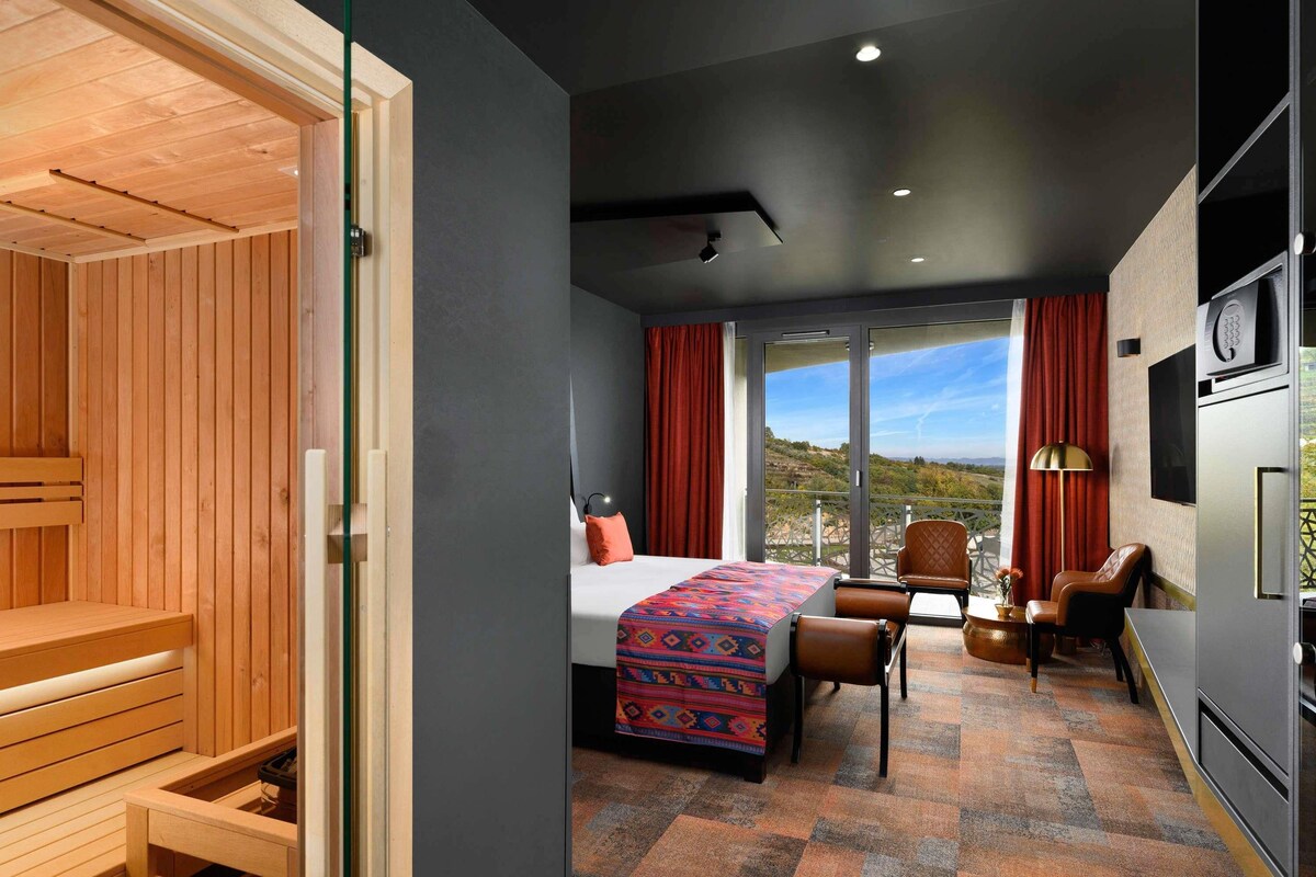 A modern hotel room with a wooden sauna, a bed with a colorful blanket, a desk with chairs, and a view of the outdoors.