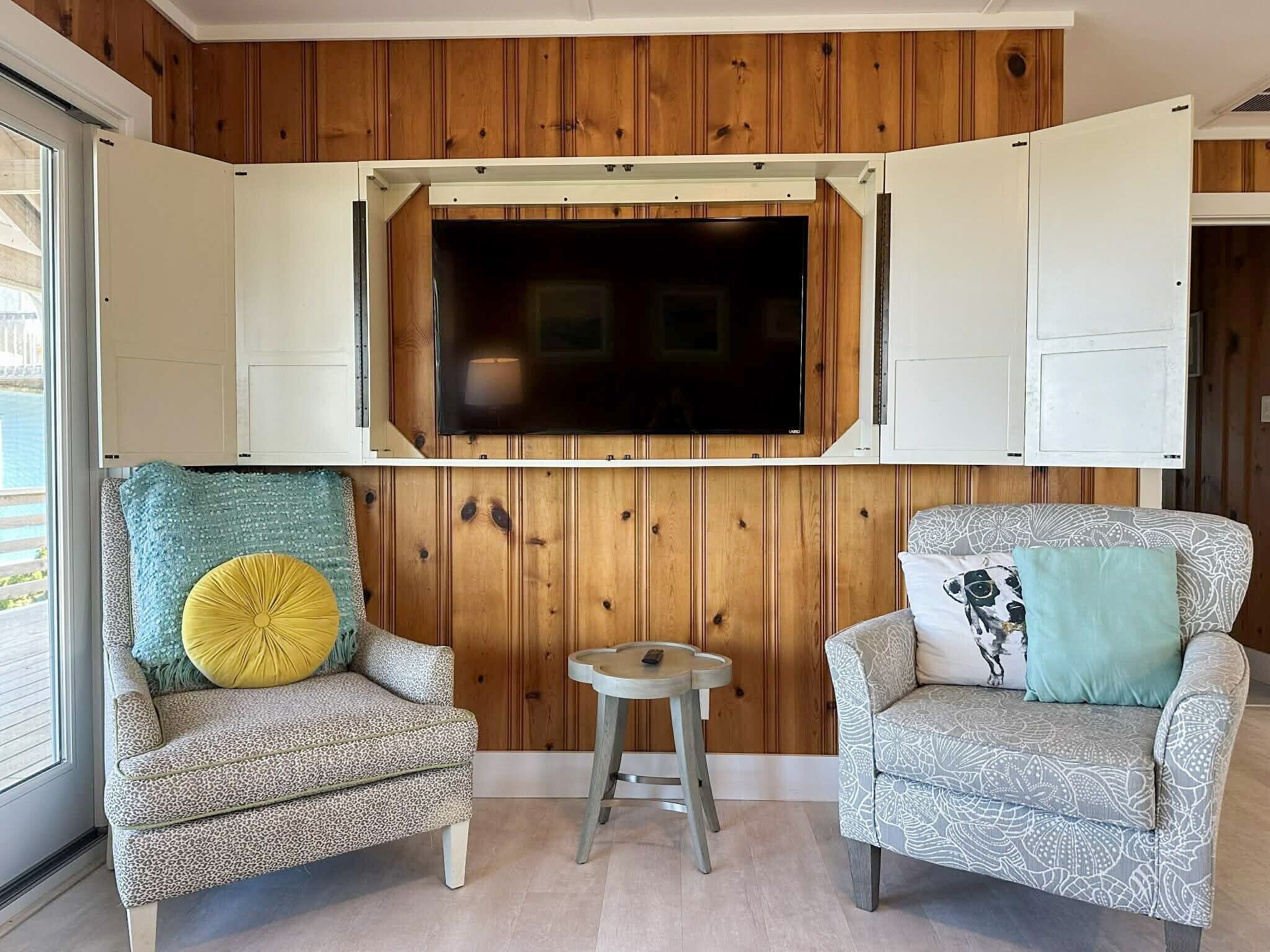 Una habitación con paneles de madera, un televisor montado, dos sillones estampados y una pequeña mesa redonda.