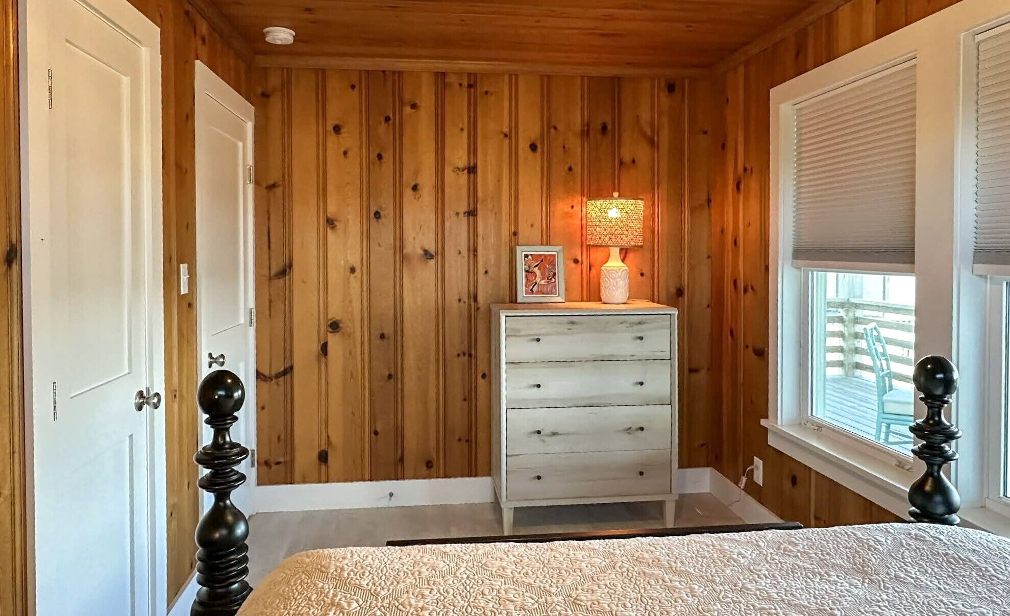 A bedroom with wooden walls, a white dresser, a bed with a patterned bedspread, a window with blinds, and a lamp on the dresser.