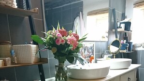 Une salle de bain avec un meuble-lavabo blanc, un miroir rond et un vase de fleurs. On y trouve des paniers sur des étagères et un sol à motifs.
