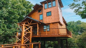 A multi-level wooden cabin with a staircase leading to the upper floor, surrounded by dense forest. - VIEWS! Luxury Treehouse with Hot Tub - Sleeps 6 (Millersburg)