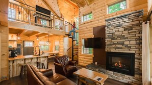A log cabin interior with a stone fireplace, leather sofa, and a kitchen area with a bar. - VIEWS! Luxury Treehouse with Hot Tub - Sleeps 6 (Millersburg)