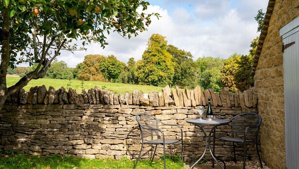 Un mur de pierre, une table avec des chaises et une bouteille de vin, le tout dans un cadre de jardin.