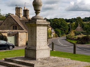 Un monument commémoratif en pierre portant des inscriptions, une maison en pierre avec une cheminée et une route qui serpente à travers un paysage verdoyant.