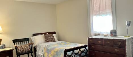 A bedroom with a bed, a wooden dresser, a chair, a nightstand, and a window with curtains.