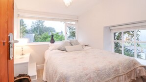 Ein Schlafzimmer mit Bett, Nachttisch, Lampe, Fenster und Blick auf Grünfläche.