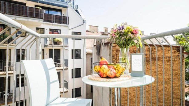 Un balcon avec une table ronde en verre, une chaise en métal, un panier de fruits et un vase rempli de fleurs.