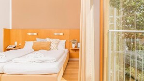 Une chambre d’hôtel avec une tête de lit en bois, un lit double avec du linge de lit blanc, une table de chevet avec un téléphone et une vue sur la verdure par la fenêtre.