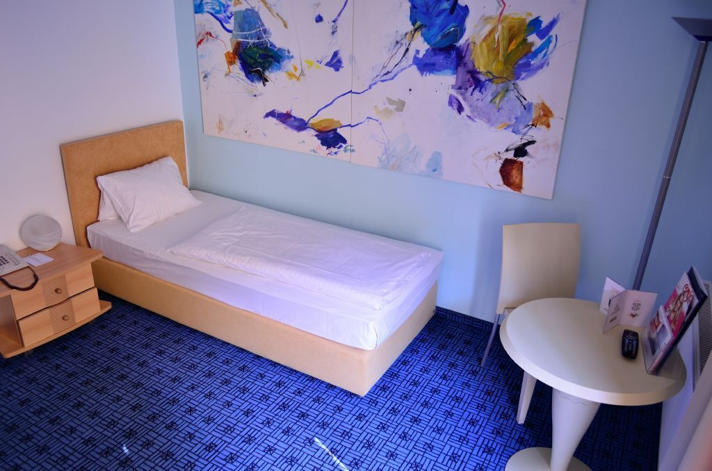 A single bed with a wooden headboard, a bedside table with a lamp, a small round table with a chair, and a large abstract painting on the wall.