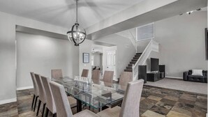 A spacious dining area with a glass table and cushioned chairs, a staircase leading to another level, and a modern chandelier. - Spacious 5 BR, 3.5 BA, 4000+sf, WiFi, Pool,Hot Tub (Gilbert)