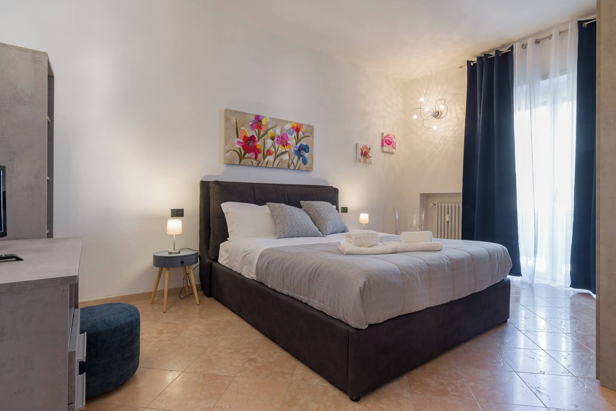 A modern bedroom with a large bed, two bedside tables, a nightstand, a chair, and artwork on the walls.