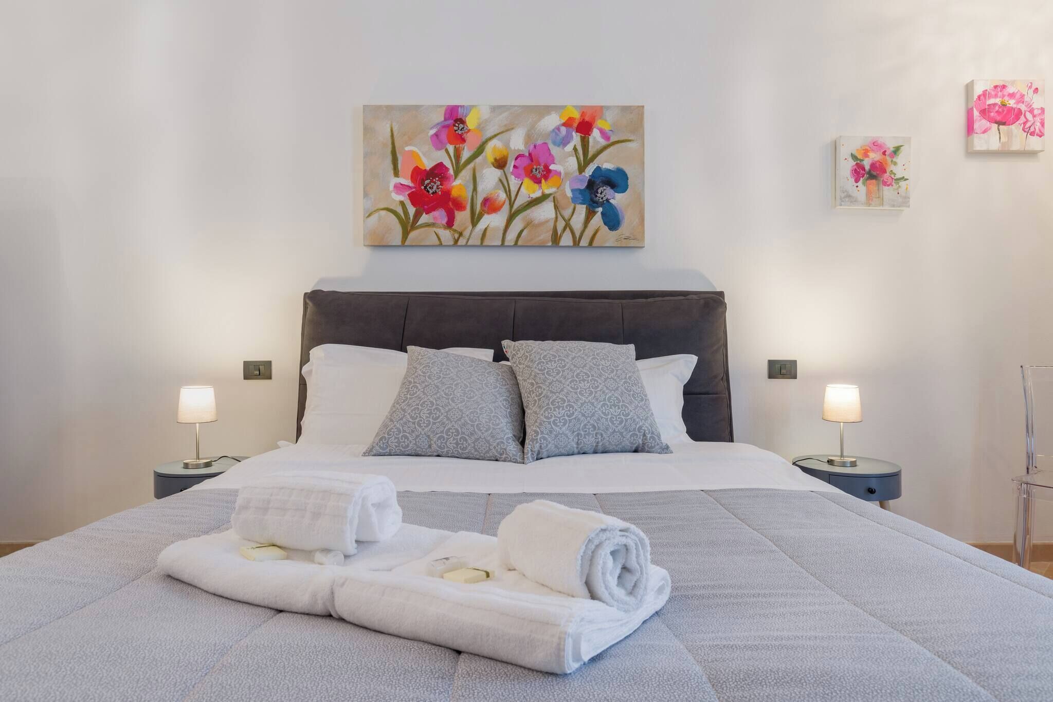 A neatly made bed with a quilt, pillows, and folded towels. There are two bedside tables with lamps and a painting above the bed.