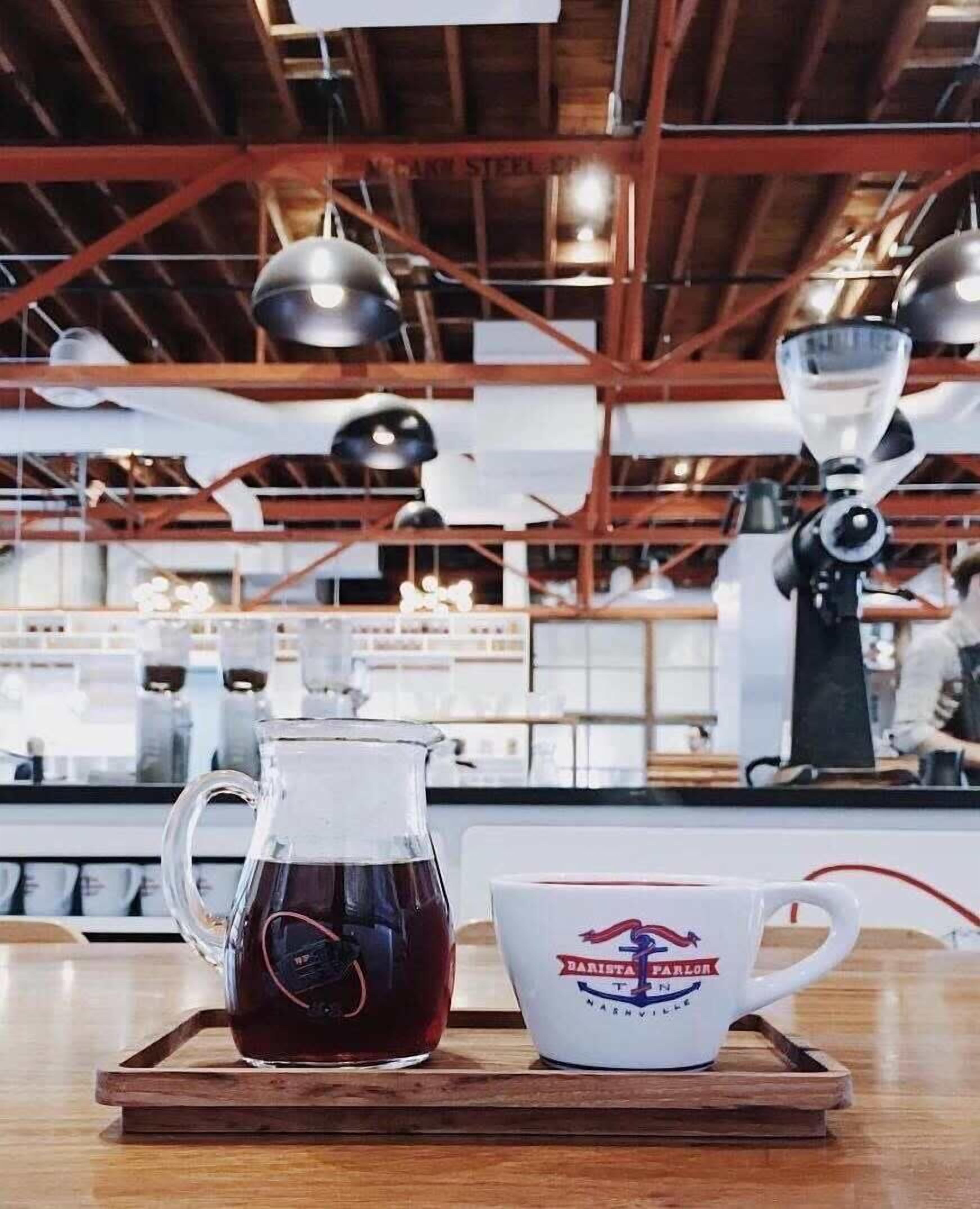 A coffee setup with a pitcher and mug on a wooden tray, in a cafe with visible industrial-style ceiling and lighting fixtures.