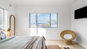 Ein Schlafzimmer mit einem Bett, einem Fenster mit Jalousien, einem an der Wand befestigten Fernseher und einem Korbstuhl.