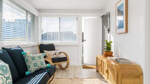 A cozy room with a dark blue sofa, patterned cushions, a wicker chair, a small wooden side table with a plant, and a view of the city through large windows with blinds. - Bella BAY 2 Upstairs - Prime Location - Holiday Yamba (Yamba)