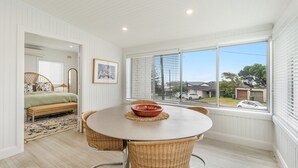 A room with a round table, wicker chairs, a bed with a green bedspread, a framed picture on the wall, and a view of the neighborhood through large windows. - Bella BAY 2 Upstairs - Prime Location - Holiday Yamba (Yamba)