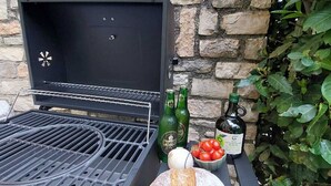 Una parrilla con un muro de piedra de fondo, una tabla de madera con una hogaza de pan, y botellas de aceite de oliva y cerveza.