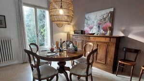 Un área de comedor con una mesa redonda, cuatro sillas, una lámpara colgante de mimbre, un cuadro en la pared y una ventana con cortinas.