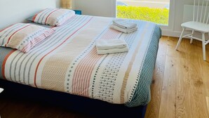 A bedroom with a bed, a chair, a window with a view of the beach, and a hanging light fixture. - La Grande Plage - Three Bedroom Apartment, Sleeps 5 (Saint-Cast-le-Guildo)
