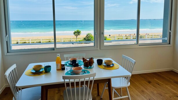 A dining area with a table set for two, overlooking a beach with a clear view of the ocean and a palm tree. - La Grande Plage - Three Bedroom Apartment, Sleeps 5 (Saint-Cast-le-Guildo)