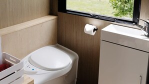A compact bathroom with a toilet, a small sink, and a window offering a view of a green landscape. - Rural Delight by Tiny Away (Ohaupo)