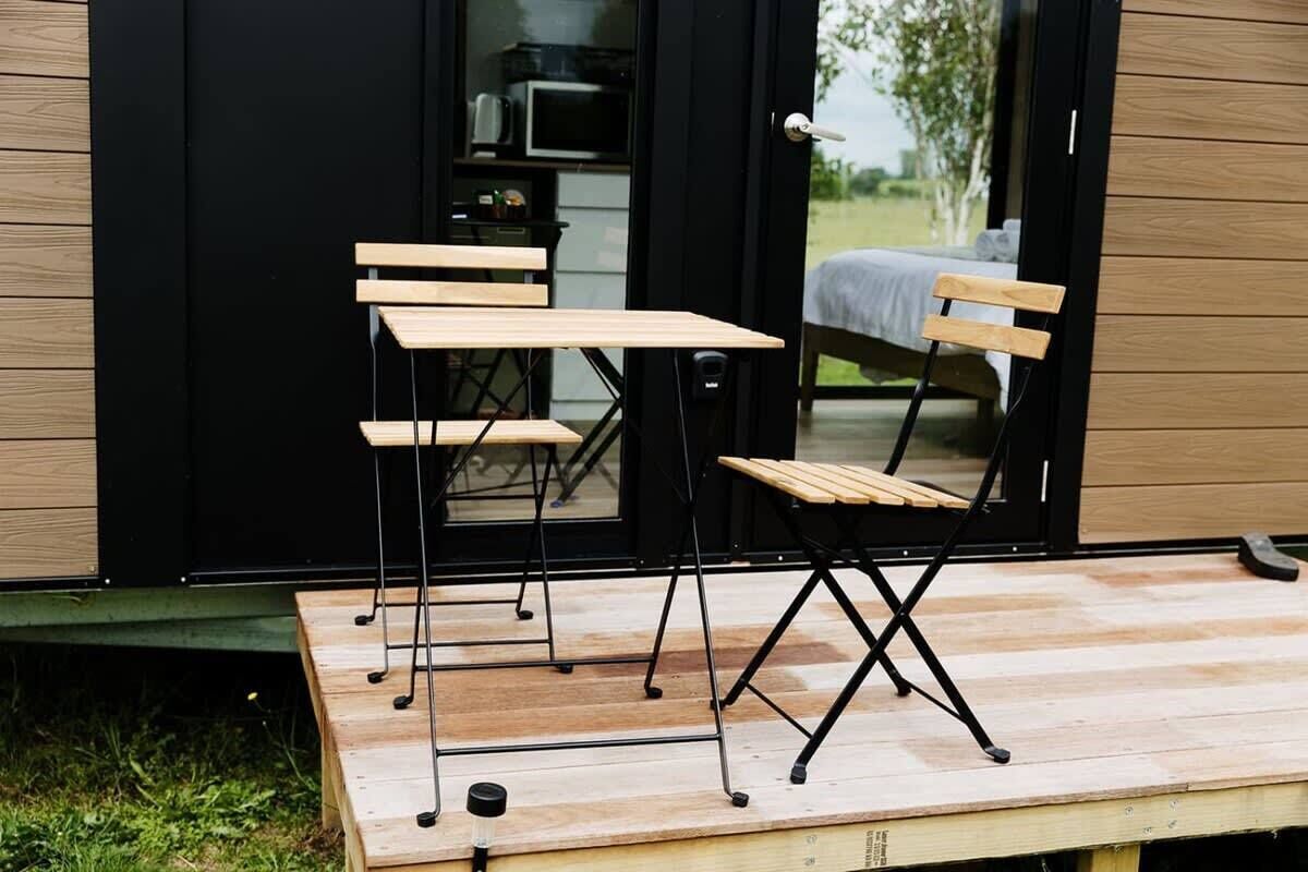 Ein kleiner Sitzbereich im Freien mit Klapptischen und -stühlen, ein Tisch mit einer Tasse und Blick in ein Schlafzimmer durch eine offene Tür.