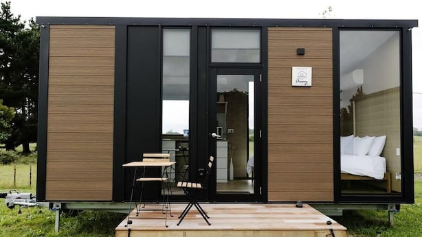 A modern, compact cabin with a wooden exterior, a glass door entrance, a small outdoor seating area, and a visible bedroom with a bed and white linens. - Rural Delight by Tiny Away (Ohaupo)