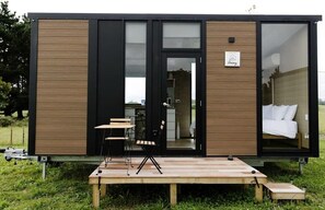Una cabaña moderna y compacta con fachada de madera, puerta de cristal, un pequeño espacio de estar al aire libre y un dormitorio visible con cama y ropa de cama blanca.