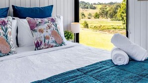 A bedroom with a large window, a bed with floral pillows, a blue rug, and a rolled white towel.