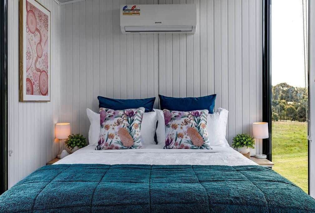 Ein Schlafzimmer mit einem Bett, zwei Kissen, einem Nachttisch mit Lampe und einem Fenster mit Blick auf Grünflächen.