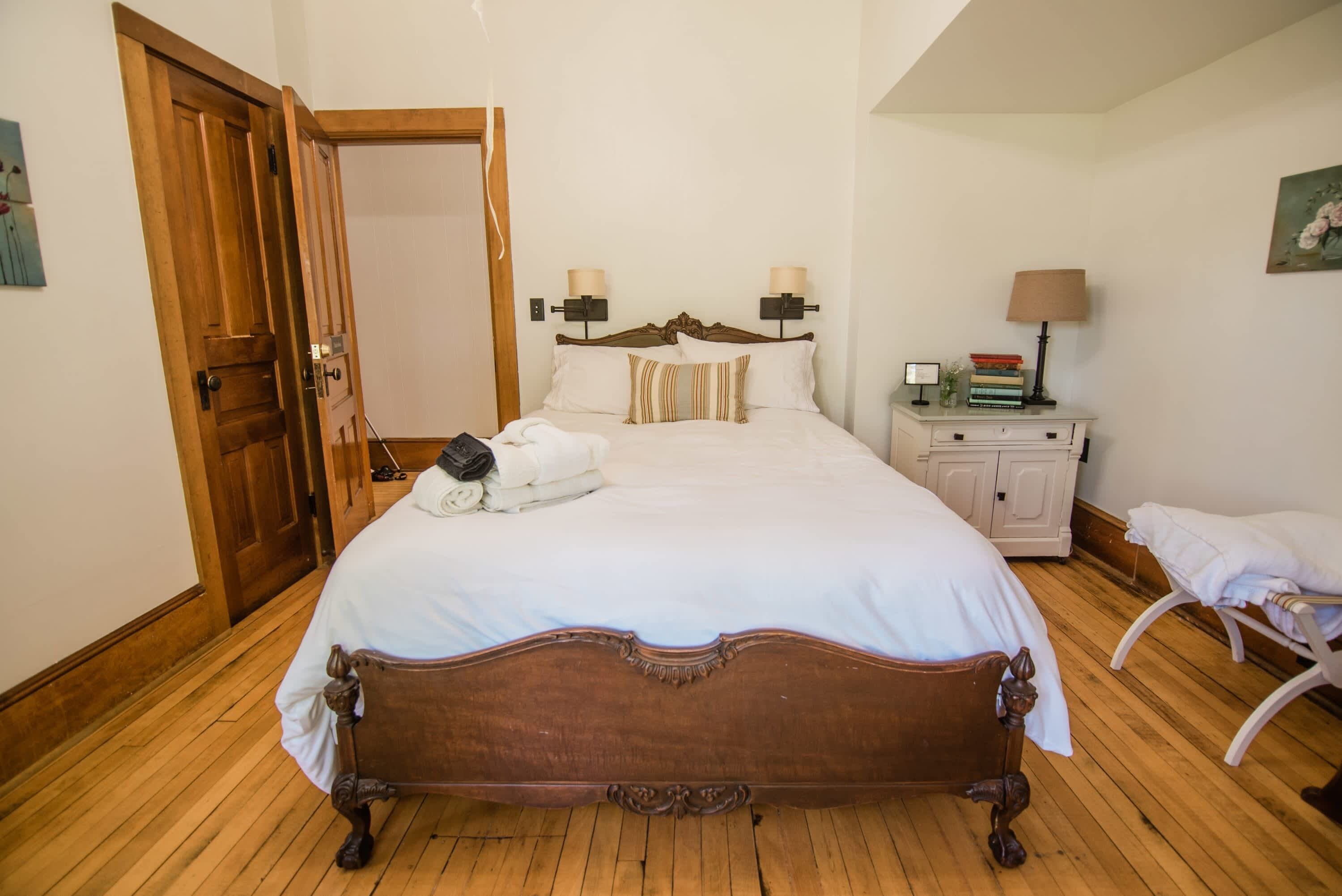 A neatly made bed with a wooden headboard, a bedside table with a lamp, and a chair with a towel.