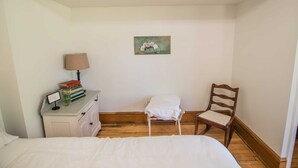 Une chambre à coucher avec un lit, une table de chevet sur laquelle se trouvent des livres et une lampe, une chaise et un tableau accroché au mur.