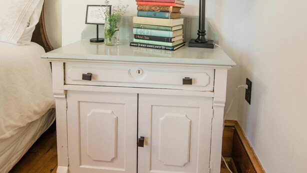 Une table de chevet blanche, avec une lampe, une pile de livres et un petit vase avec des fleurs.