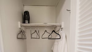 Un petit dressing avec une armoire en bois, une robe blanche suspendue et une étagère sur laquelle se trouvent un ventilateur et des cintres.