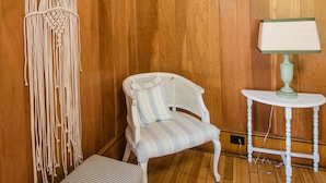 A room with wooden panel walls, a white upholstered chair, a small white table with a lamp, and a macramé wall hanging.