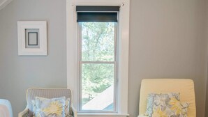 Ein Zimmer mit Holzboden, einem Fenster mit Blick auf Bäume, einem weißen Sessel mit Kissen und einem gelben Sessel mit Kissen.