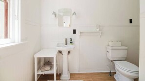 Une petite salle de bain blanche, avec un lavabo sur colonne, un miroir, des toilettes et une étagère au-dessus des toilettes.