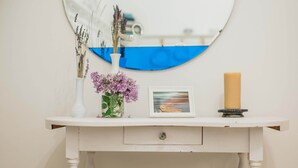 Une coiffeuse en bois blanc avec un miroir rond, un petit vase blanc contenant des fleurs violettes, un tableau encadré et un chandelier.