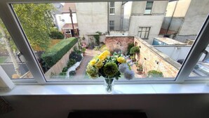 Una ventana con vista a un patio con pared de ladrillo, un pequeño jardín y un florero con flores en el alféizar.