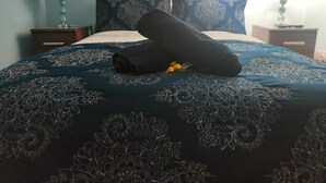 Una cama cuidadosamente tendida con un edredón azul oscuro con estampado floral y almohadas a juego, una toalla enrollada y una pequeña flor amarilla sobre la cama.