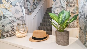 A room with a white wall and a patterned wallpaper. There is a potted plant, a glass jar, and a straw hat on a white surface. - Eller Close House (Ambleside)