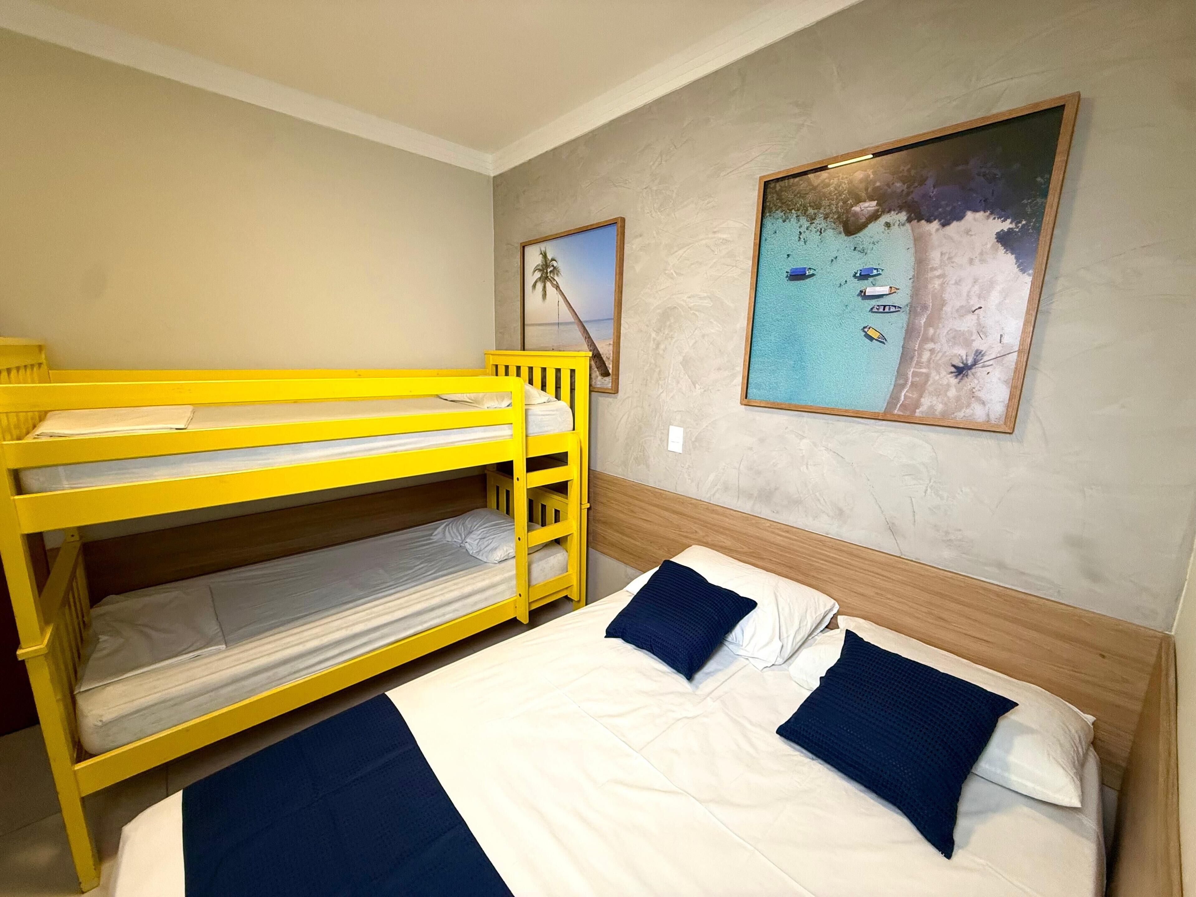 A room with bunk beds and a single bed, a painting of a beach scene, and a plain wall.