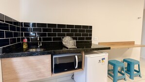 A compact kitchen with black tiled backsplash, a stainless steel sink, a microwave, and a small refrigerator. There are two blue stools and a wooden shelf above the counter. - Marmeu Tropical Ltda (Guarujá)