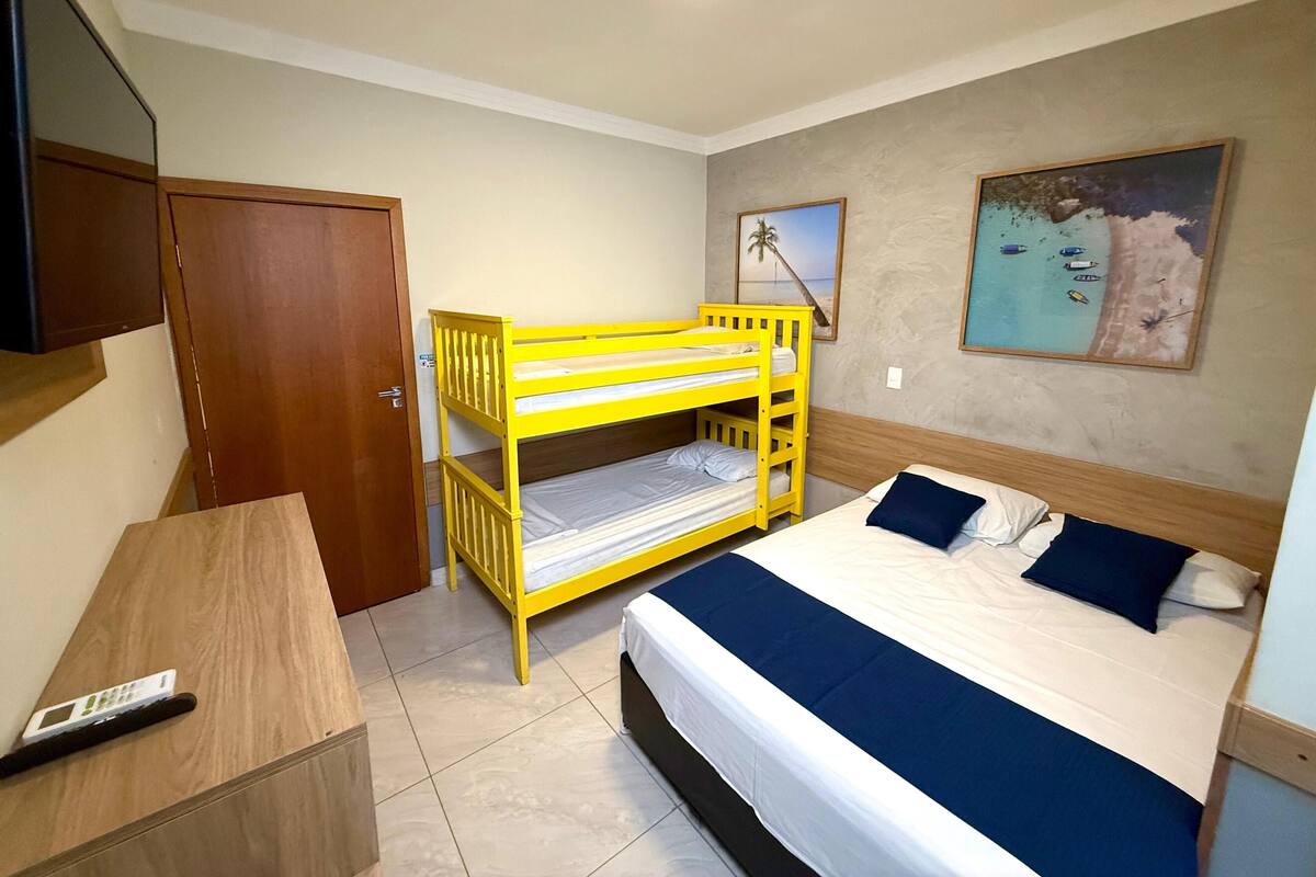 A bedroom with a bunk bed, a single bed, a wooden desk, a television, and a beach-themed wall decoration.