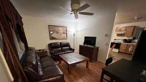 Una sala de estar con un sofá de cuero, una mesa de centro, un televisor sobre un mueble de madera y un ventilador de techo.